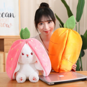 A person is shown partially, with only the face visible, smiling behind two large plush toys resembling a strawberry and a peach, placed on top of a closed laptop on a wooden table. A motivational quote on the wall and a houseplant in the background contribute to a cozy atmosphere.