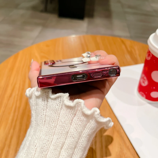 A person wearing a white knit glove with a ruffled edge is holding a maroon smartphone with a reflective case showing a cartoon character. In the background, there's a red cup with white polka dots and a white plate on a wooden table surface.